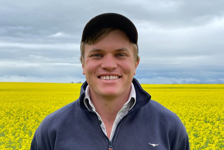 man in a cap smiling at camera with a field of yelloe canola behind him 