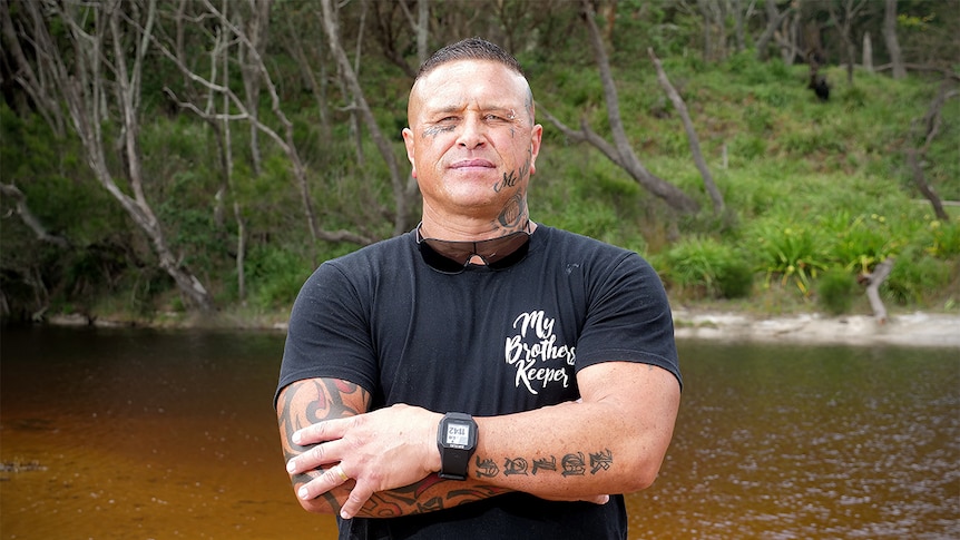 James Williams stands in a creek with his arms folded, wearing a black t-shirt and shorts.