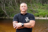 James Williams stands in a creek with his arms folded, wearing a black t-shirt and shorts.