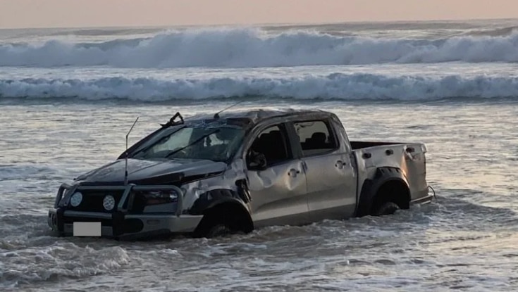 A dented black car with water up to its wheels on a beach. 
