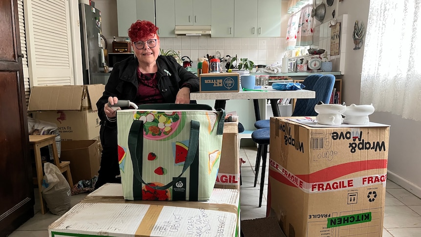 An older, bespectacled woman with dyed hair stands amid packing boxes in a kitchen.