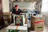 An older, bespectacled woman with dyed hair stands amid packing boxes in a kitchen.