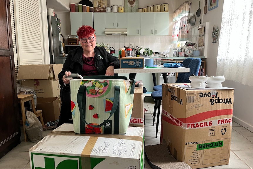 Lesley sits at her kitchen table surrounded by boxes for her move she has bright red short hair and a walking stick
