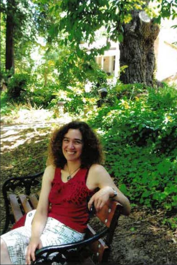 Slightly grainy photo of a younger Hilary Harper, with dark curly hair, red singlet and pants, sitting on park bench, smiling.