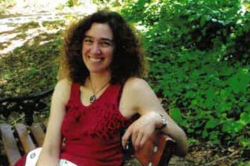 Slightly grainy photo of a younger Hilary Harper, with dark curly hair, red singlet and pants, sitting on park bench, smiling.