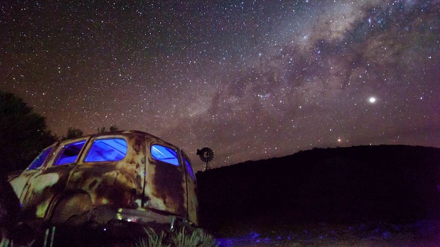 A car wreck against a backdrop of a hill and a night sky lit up with stars.
