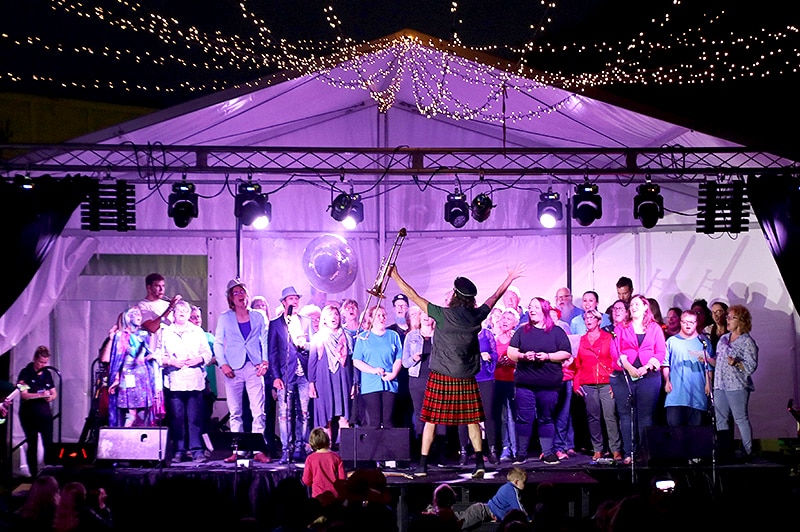 A choir sings on stage as directed by a conductor holding a trombone.