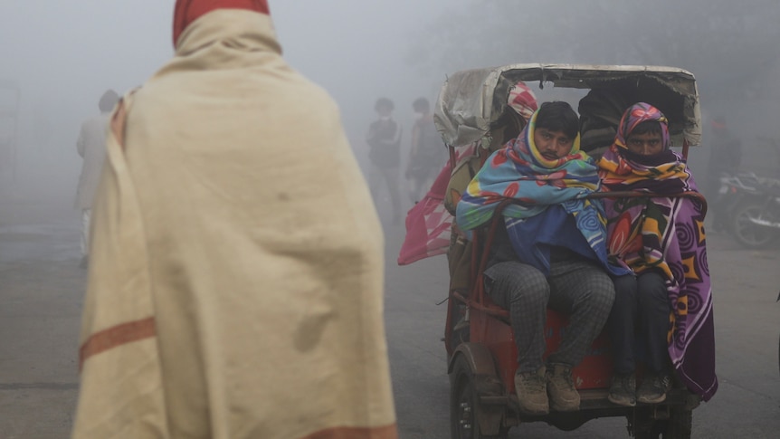 Two men bundled in colourful quilts sit on the back of a ricksaw during heavy fog.