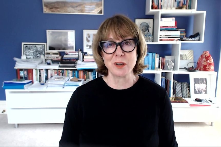 A woman with short hair with black glasses in a black shirt, sitting in an office with bookshelves behind her.