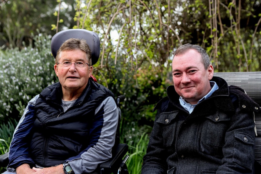 Man in blue shirt sits next to man in wheelchair wearing grey and black top with glasses, with garden behind them