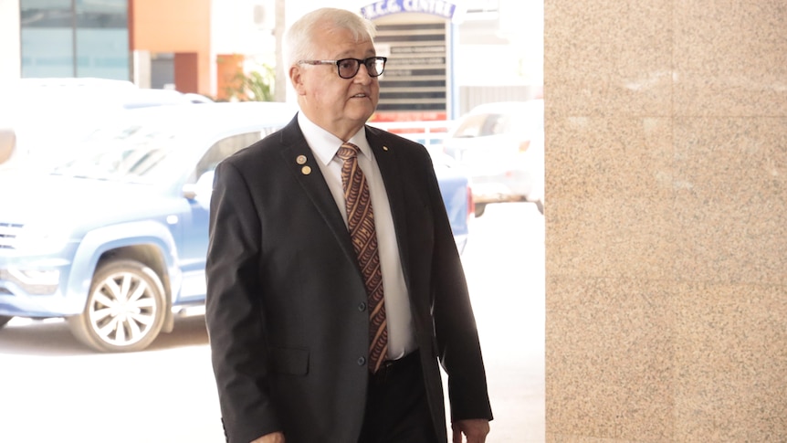 An older man with white hair and a suit and tie walking into a hotel lobby.