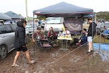 a group of people stand and sit under a covering near cars at a muddy campsite