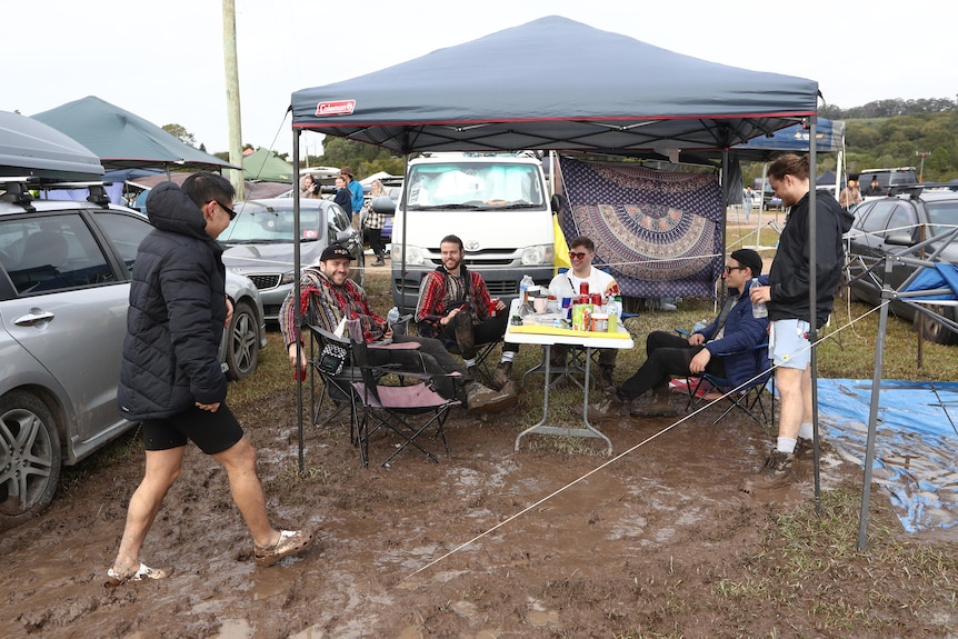 a group of people stand and sit under a covering near cars at a muddy campsite