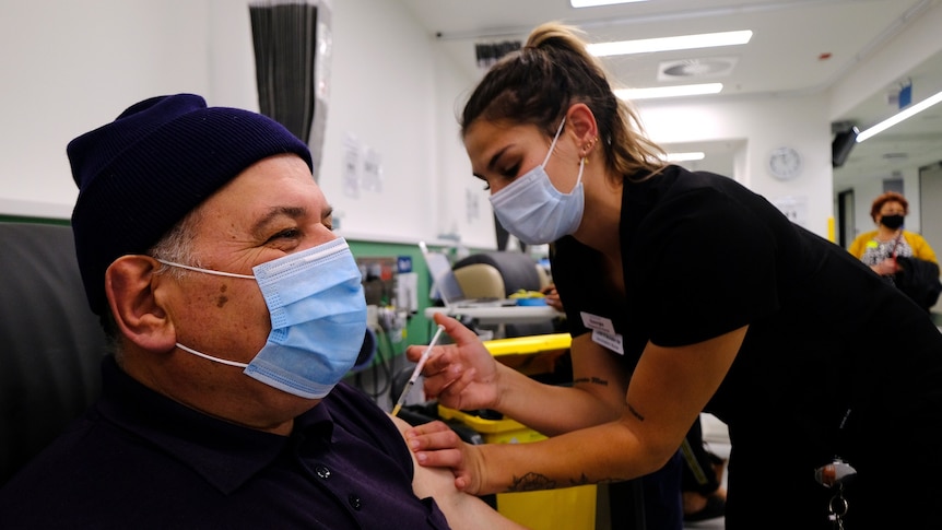 A vaccinator in a surgical mask administered a needle to a man with a mask and beanie on, smiling, in a chair.