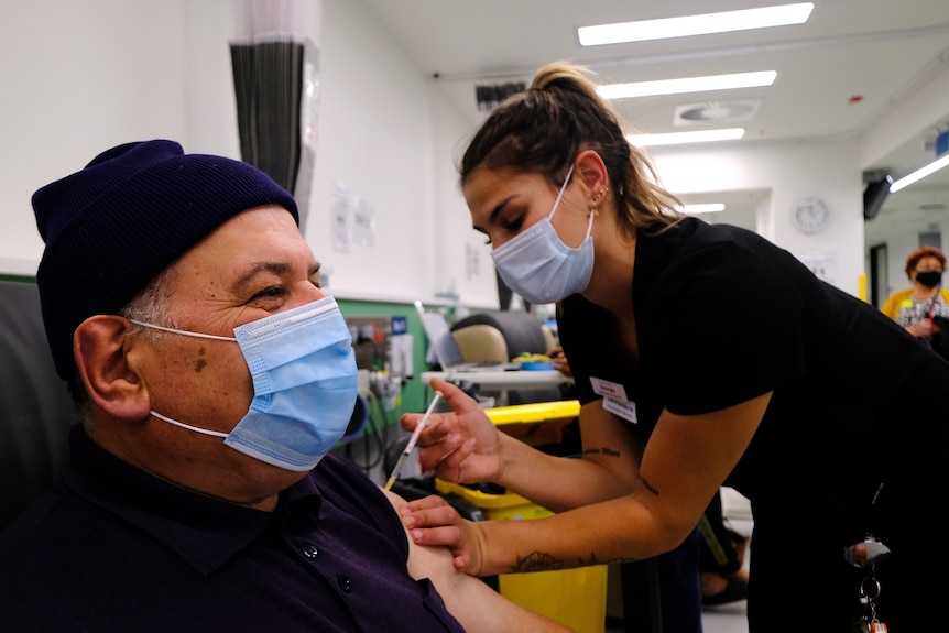 A vaccinator in a surgical mask administered a needle to a man with a mask and beanie on, smiling, in a chair.