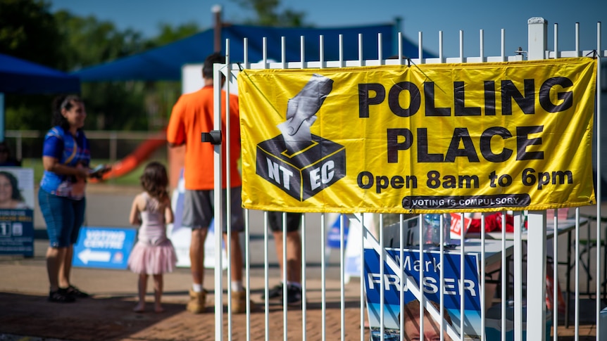 People stand inside a gate that has a bright yellow sign on it reading 'Polling place'.