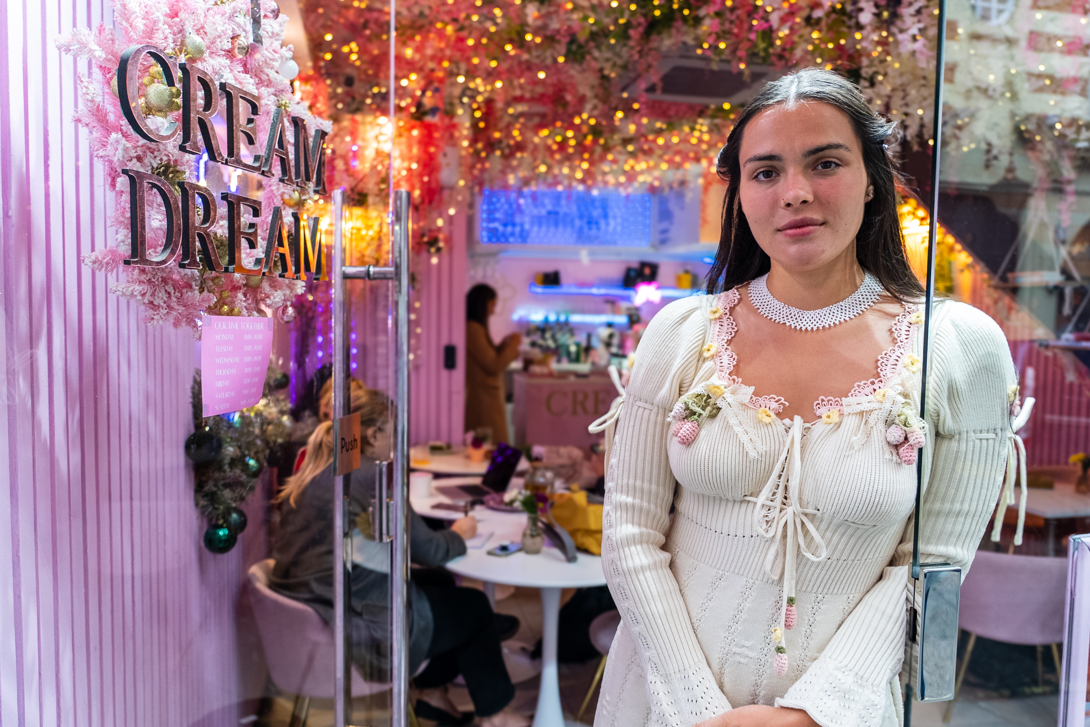 A woman in a white dress with long brown hair stands in front of a cafe door with the words 'cream dream' on it