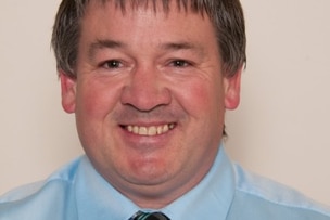 Headshot of a man with a light blue shirt and tie standing against a white wall.