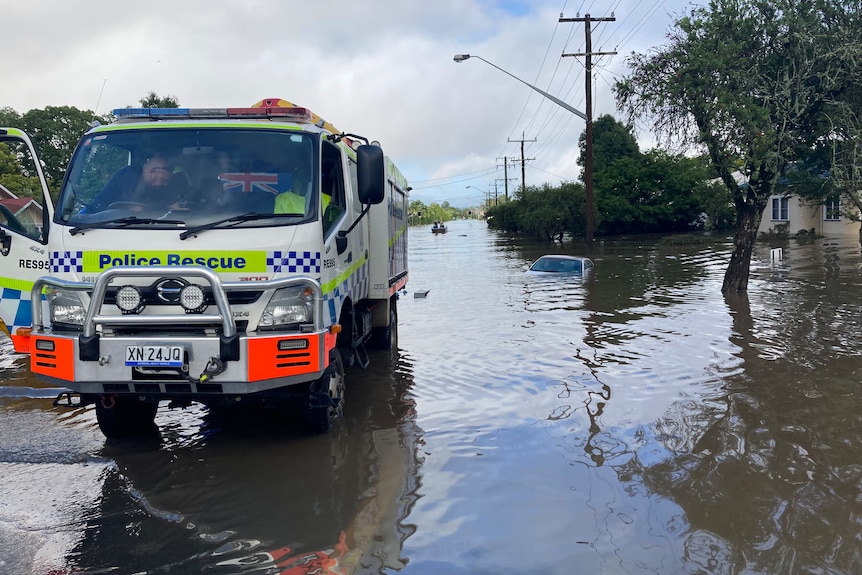 Police rescue truck parked on a road at the edge of flood waters. Submerged car in the distance