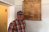 Blurred photo of an older man wearing red and black cap and check shirt stands near a near trophy board. Slight smile.