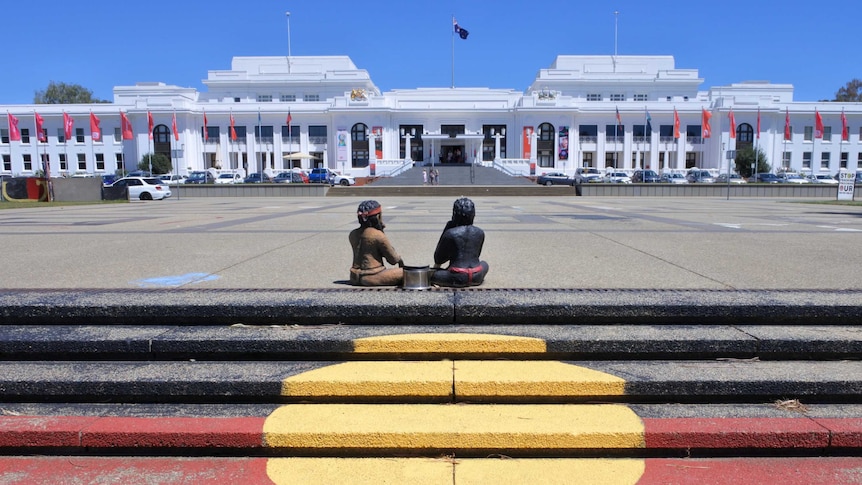 two small statues of First Nations Australians sit facing Old Parliament House on top of steps painted in the Aboriginal flag
