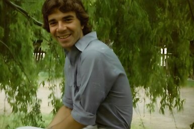 An old photograph of a young man with olive skin, floppy brown hair and a beaming smile, sitting under a willow tree.