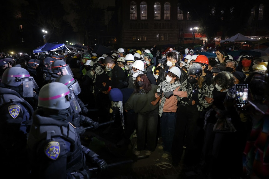 A line of police in helmets face a group of protestors in hard hats, goggles and masks
