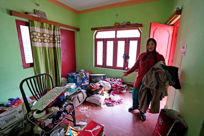 A woman gestures towards her belongings on the floor inside a house.