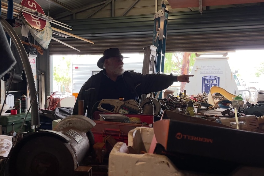 A man stands in a shed packed with items, pointing to something out of sight of the camera. 