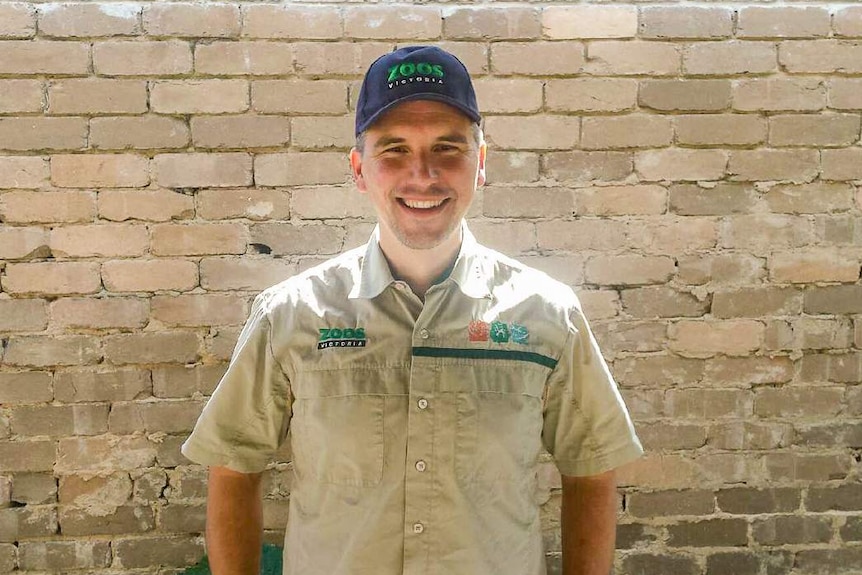 A man in a khaki shirt stands in front of a light brown brick wall.