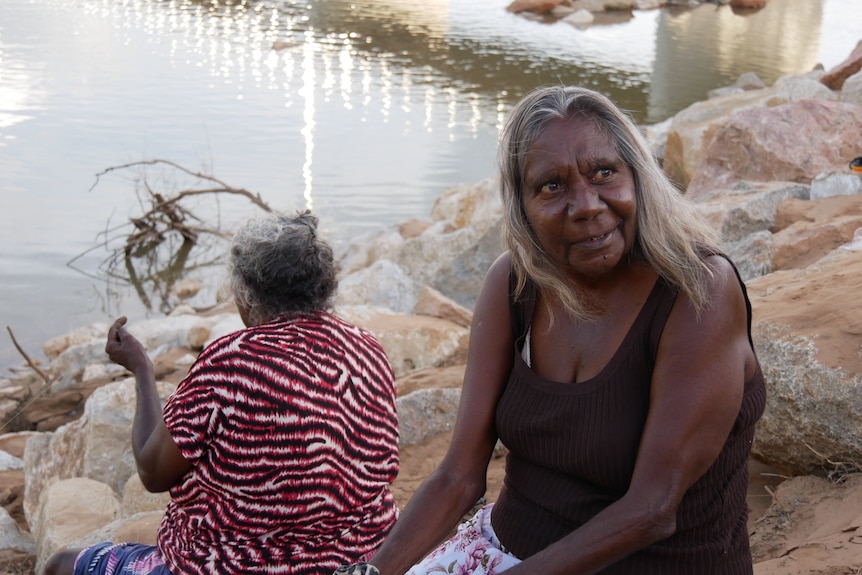 Two women sitting on rocks next to a river, as one of the women is fishing.