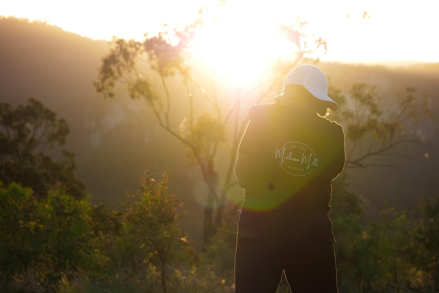 Melissa standing, camera to her face, sun setting behind her, lens flare, mountains and trees.