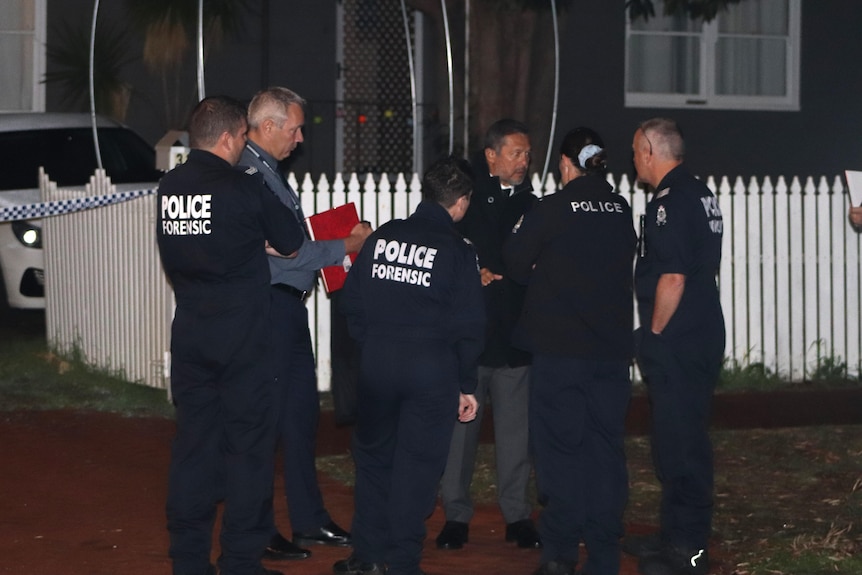 A group of police forensic officers and plain-clothes officers stand in a driveway outside a house at night.