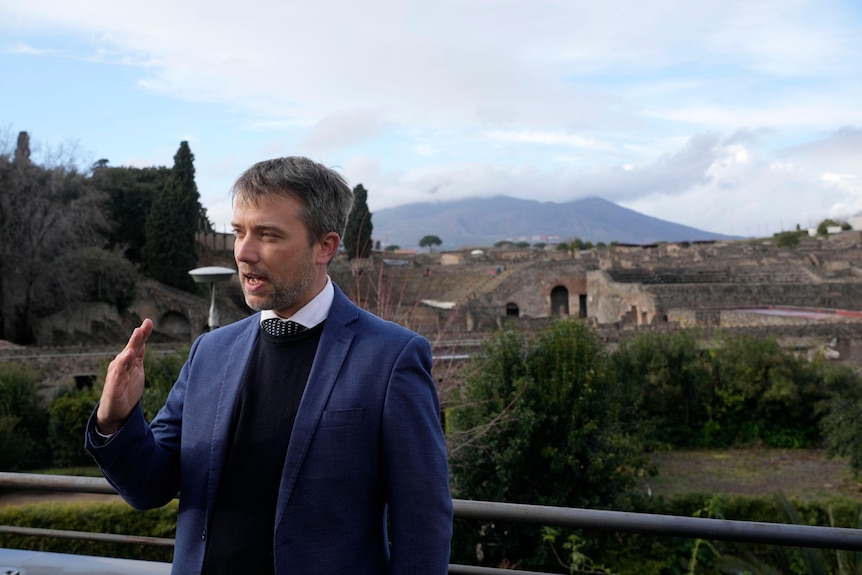 Director in a suit speaks during an interview in front of an Italian archaeological site.