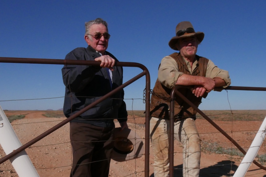 An elderly man and a man of late middle age stand against a farm fence in a dusty paddock.