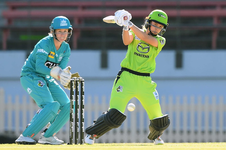A Sydney Thunder WBBL player hits out to the leg side as the Brisbane Heat wicketkeeper looks on.