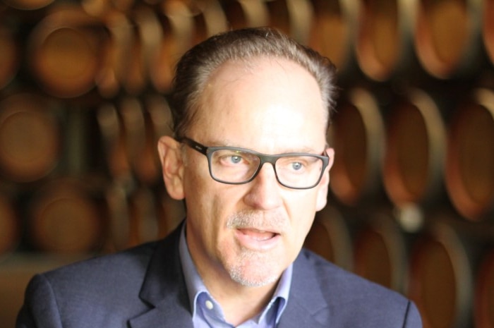 A man sits speaking, behind are are many rows of round wine barrels