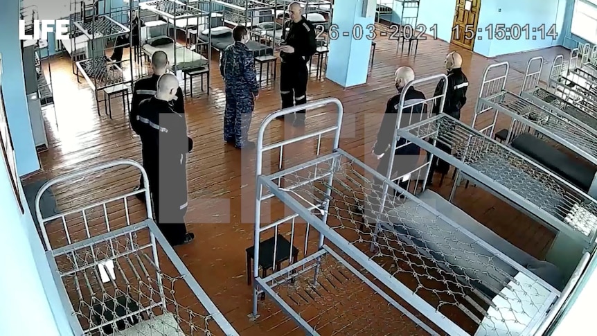 Security camera footage shows a man surrounded by guards and beds without mattresses in a prison.