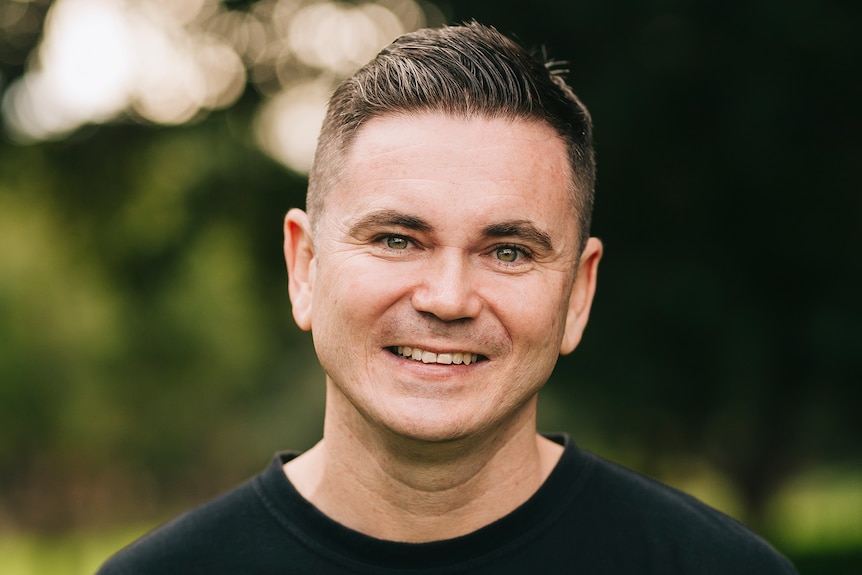 A man with short hair smiles brightly while standing outside under a tree