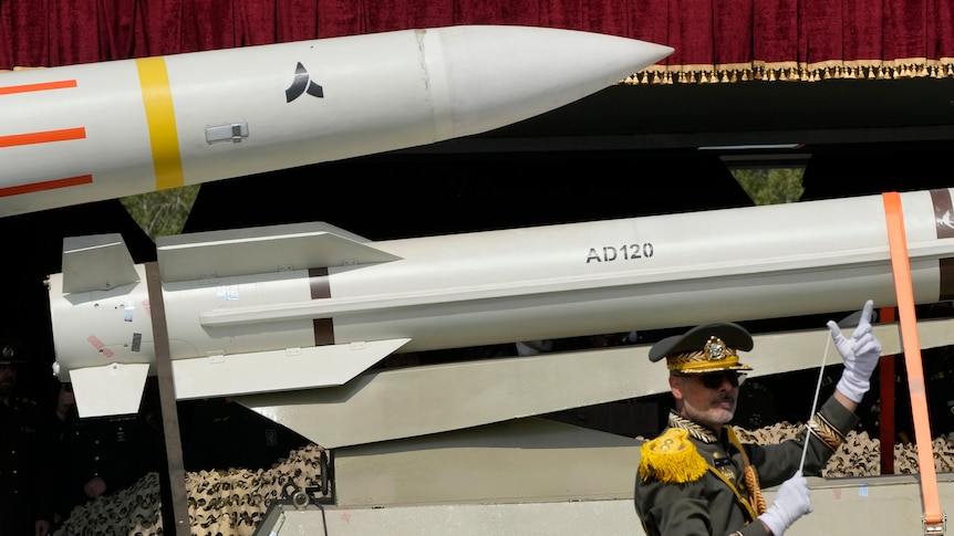 A band conductor orchestrates in front of two missiles being carried on the back of a truck