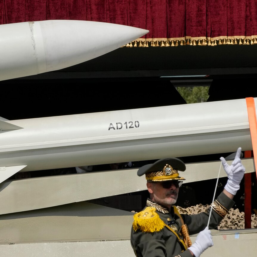 A band conductor orchestrates in front of two missiles being carried on the back of a truck