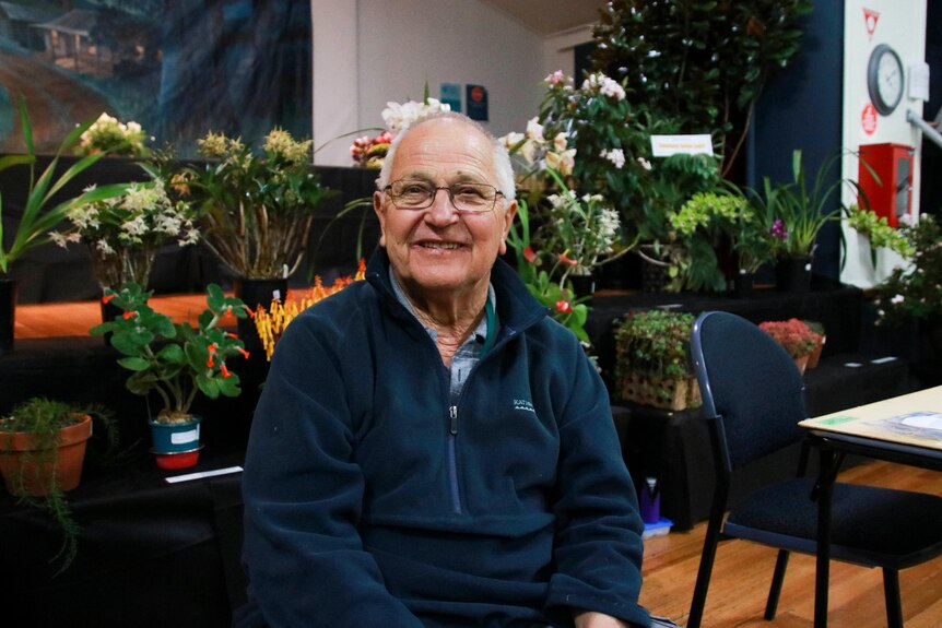 A man sitting in front of a row of floral arrangements smiles at the camera
