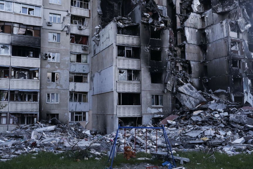 A teddy bear hangs on a swing next to a damaged building.