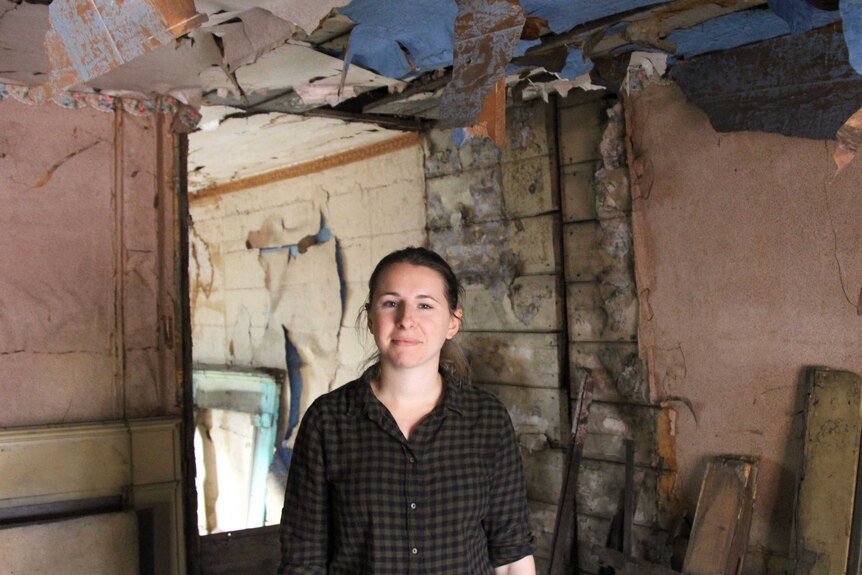 A woman stands in a dilapidated room with wallpaper peeling off the walls and ceiling.