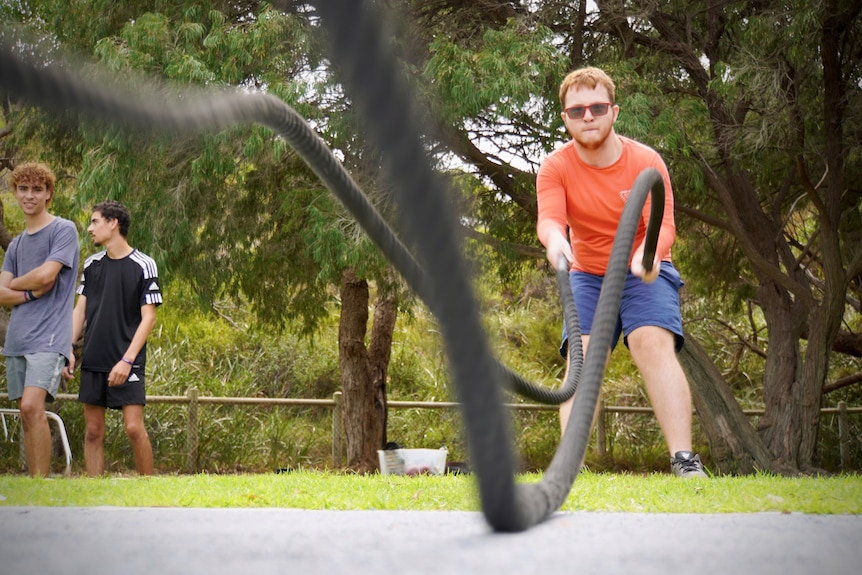 A young man in red glasses uses battle ropes during a fitness class outdoors. He is wearing an orange tee and blue shorts.