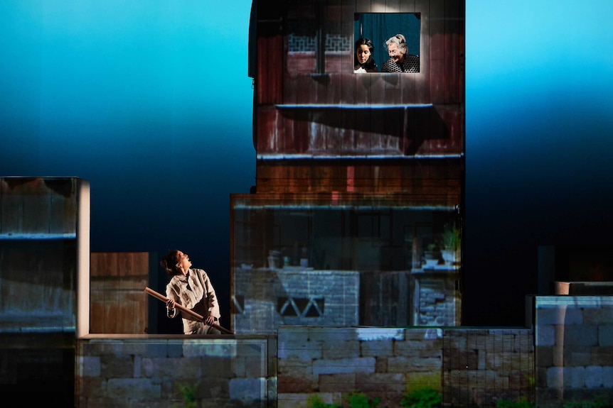A stage with a woman rowing a boat, and two other women up in a window of a ramshackle building