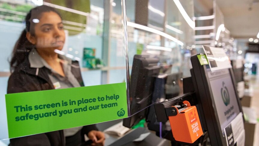 A Woolworths employee stands at a checkout behind a see-through screen