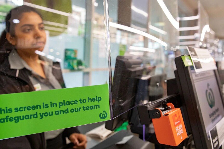 A Woolworths employee stands at a checkout behind a see-through screen