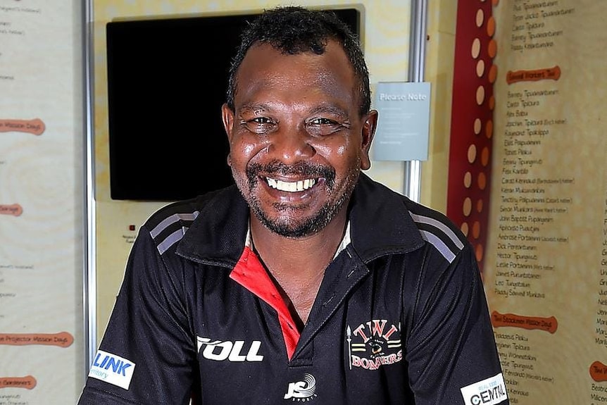 Willie Rioli Senior tersenyum ke arah kamera yang mengenakan kaos Tiwi Bombers.