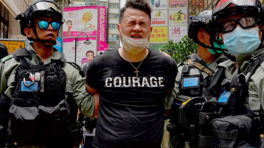 A handcuffed man wearing a black t shirt with "courage" printed across his chest squints as riot police move him.
